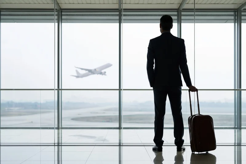 Businessman in silhouette standing at airport terminal window with suitcase, watching airplane depart