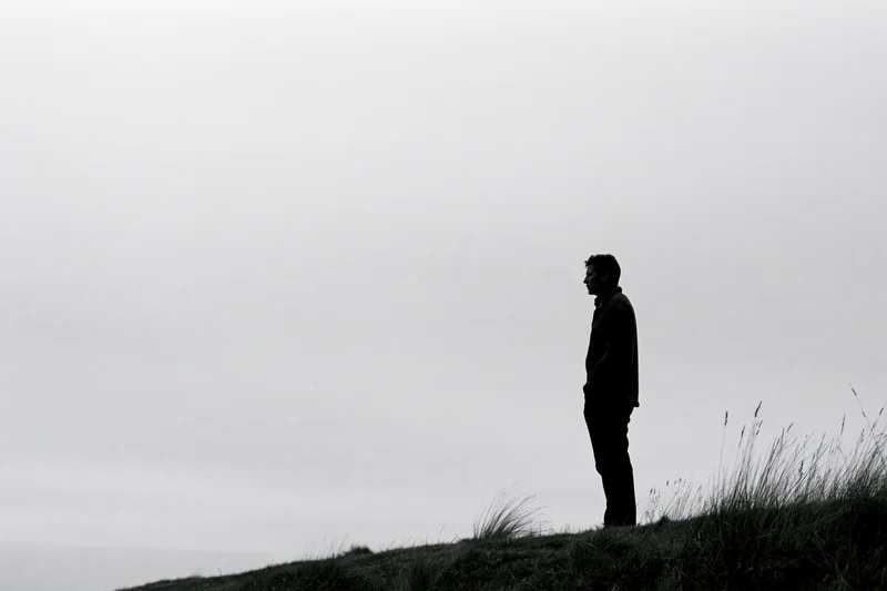 Silhouette of a man standing alone on a grassy hill under an overcast sky