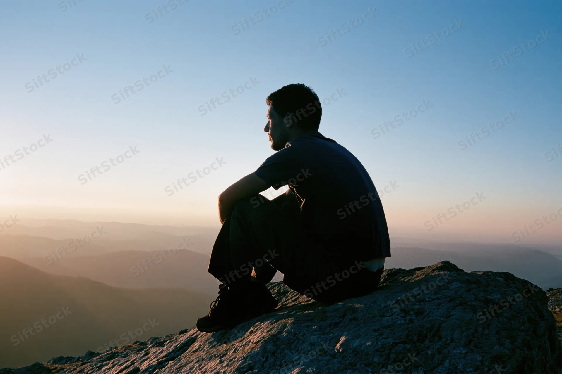 Young man sitting in silhouette on rocky mountain summit overlooking valley at sunset