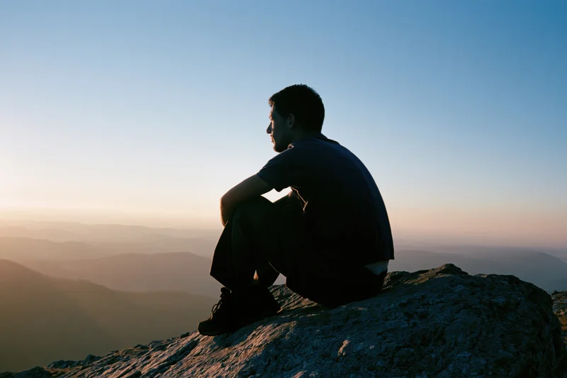 Young man sitting in silhouette on rocky mountain summit overlooking valley at sunset