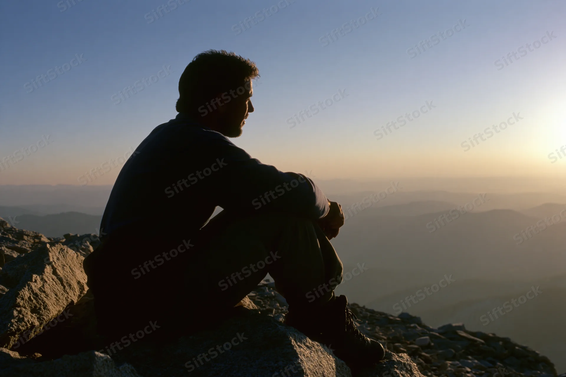 Young man in silhouette sitting on rocky mountain summit overlooking layered valleys at sunrise