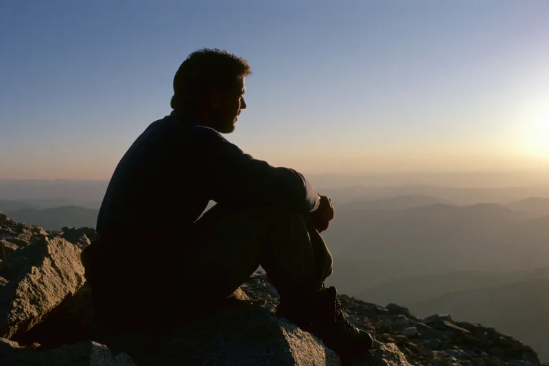 Young man in silhouette sitting on rocky mountain summit overlooking layered valleys at sunrise