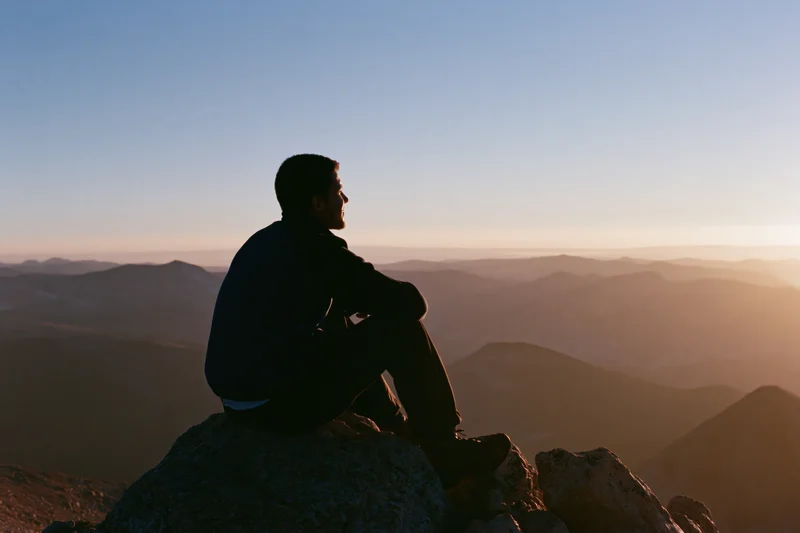 Young man sitting on rocky mountain summit overlooking layered valleys at sunset