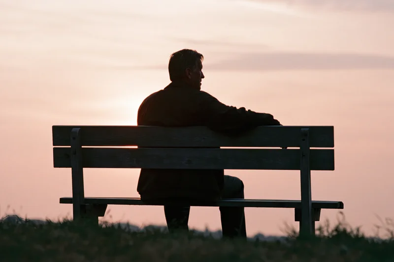 Elderly man sitting alone on wooden bench at sunset in park
