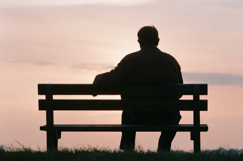 Elderly man sitting alone on a wooden park bench at sunset, back view against a soft pastel sky