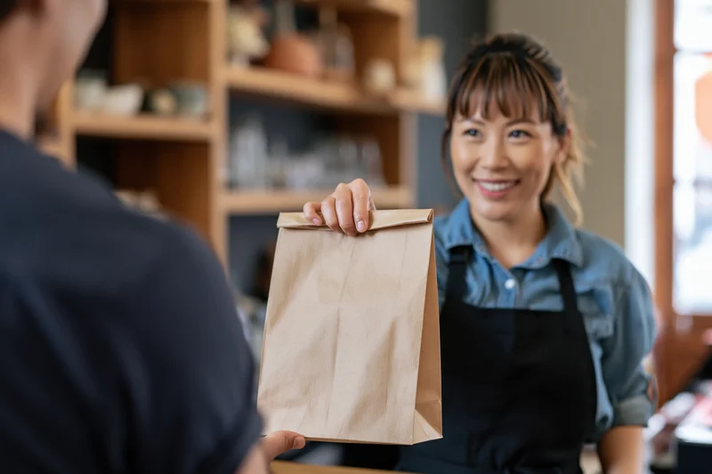 Smiling woman barista handing a brown paper bag to a customer at a cafe counter