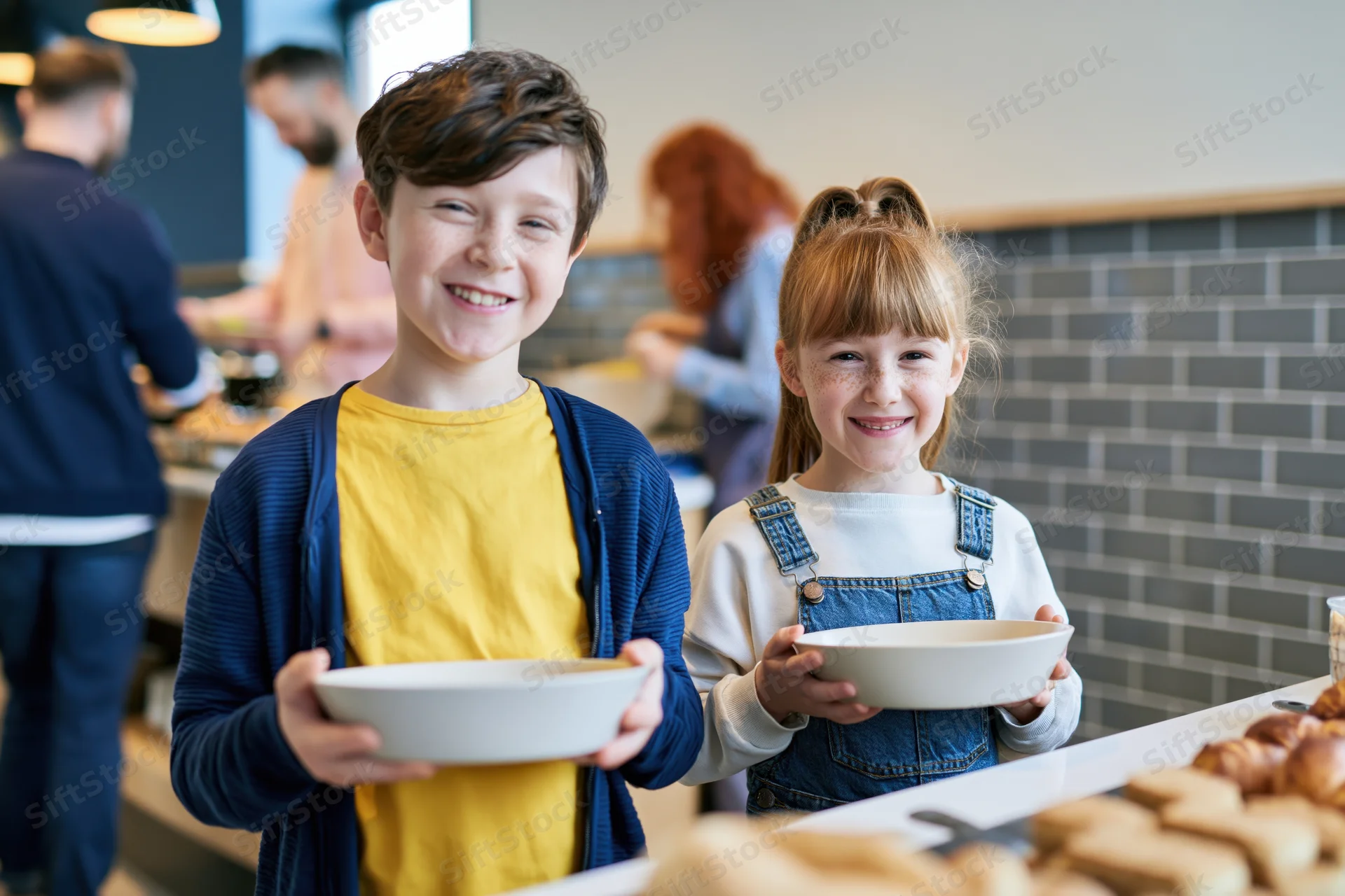 Two smiling siblings holding empty bowls at a hotel breakfast buffet