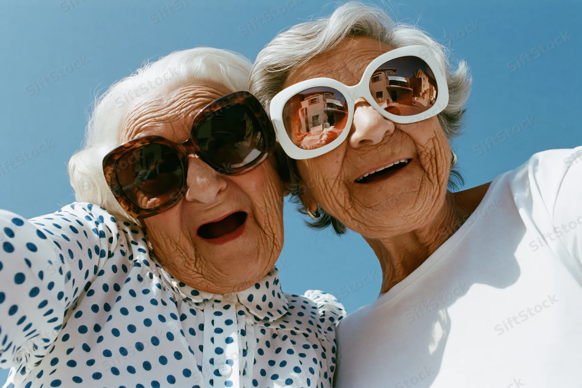 Two elderly women laughing cheek-to-cheek outdoors under a clear blue sky