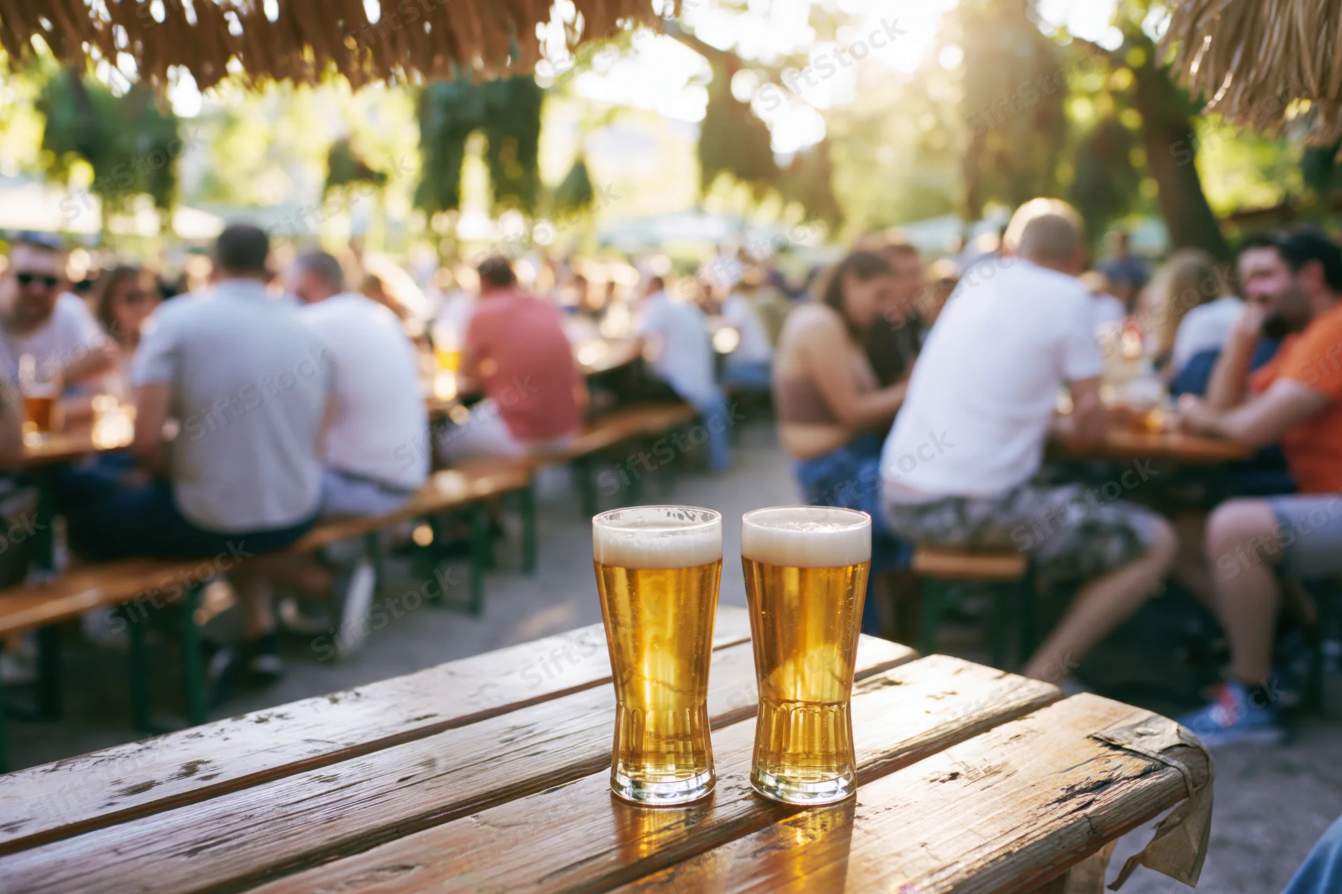 Two glasses of pale lager on a worn wooden picnic table in an outdoor garden setting with people socializing in the background under a thatched roof