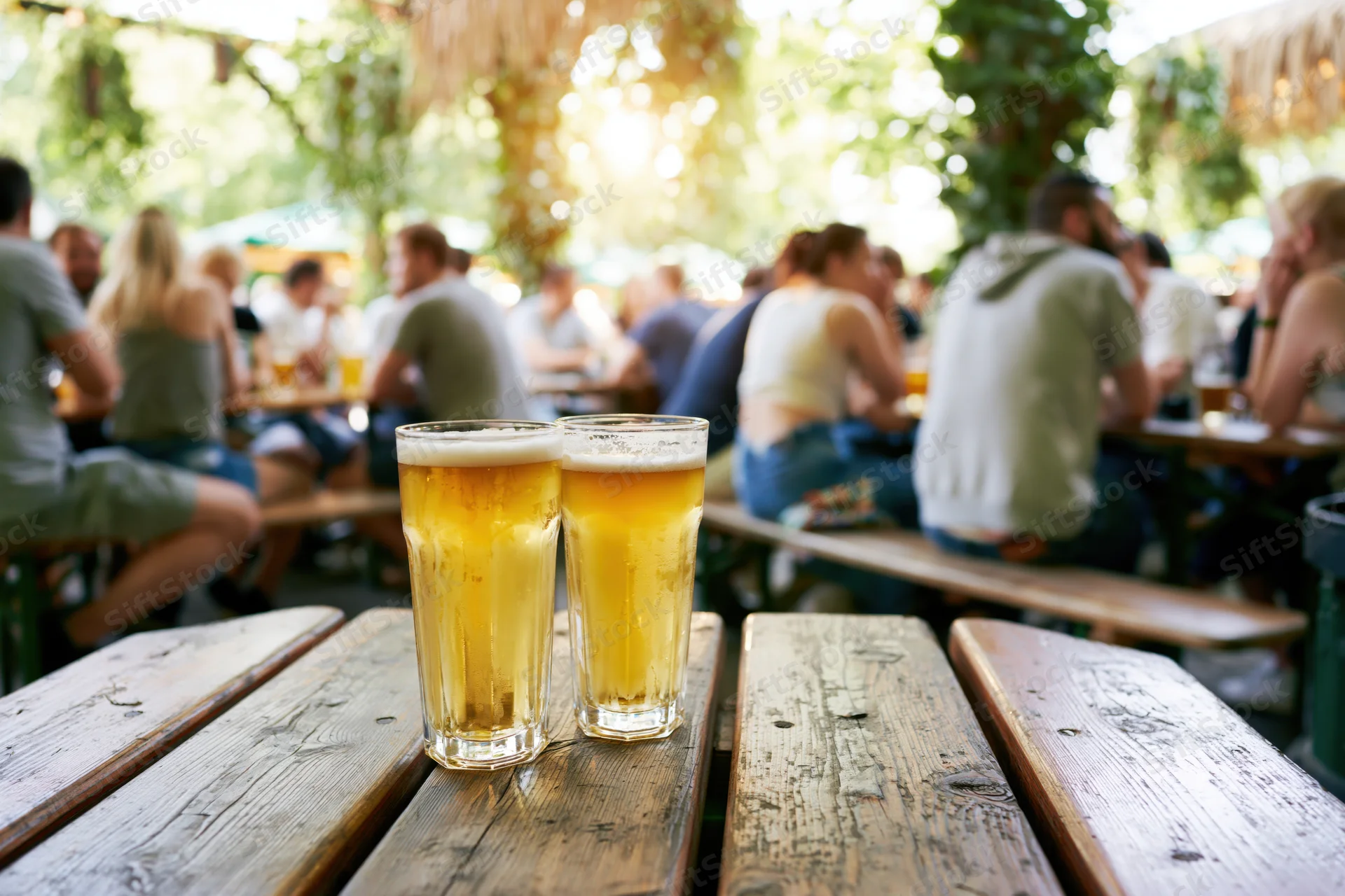 Two glasses of pale lager on a worn wooden picnic table in an outdoor garden pub setting with people socializing in the background