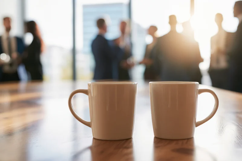 Two white ceramic coffee mugs on a wooden table in an office with blurred business people in the background during a meeting
