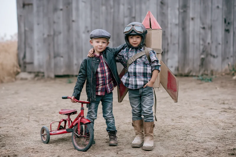 Two young boys posing together in a rural farmyard, one wearing a cardboard rocket costume and helmet, the other holding a red tricycle