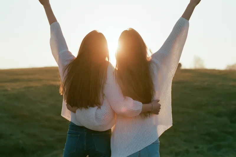Two young women embracing with arms raised toward the sun in a grassy field at sunset