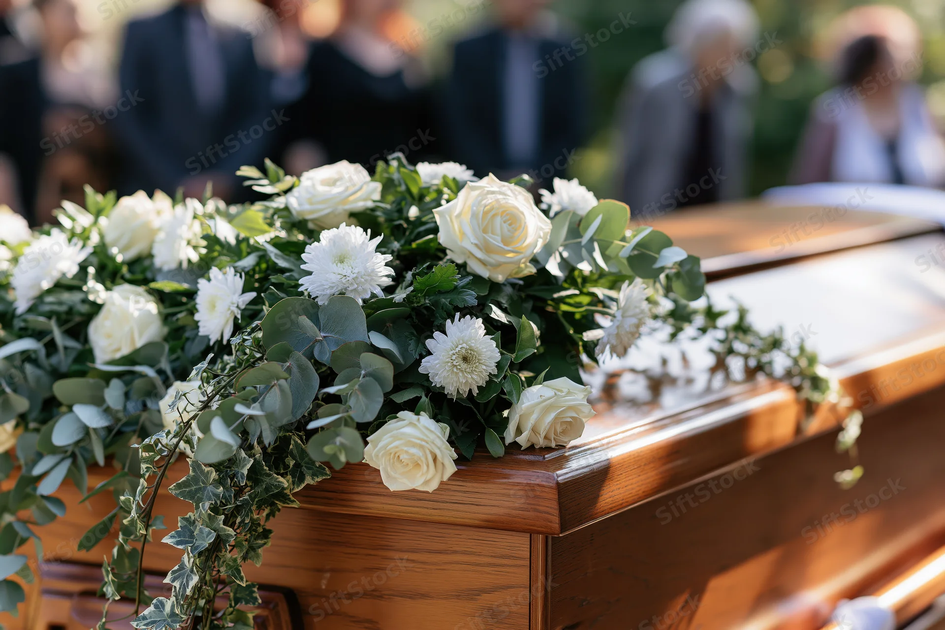 White roses and chrysanthemums draped over a wooden coffin at an outdoor funeral service