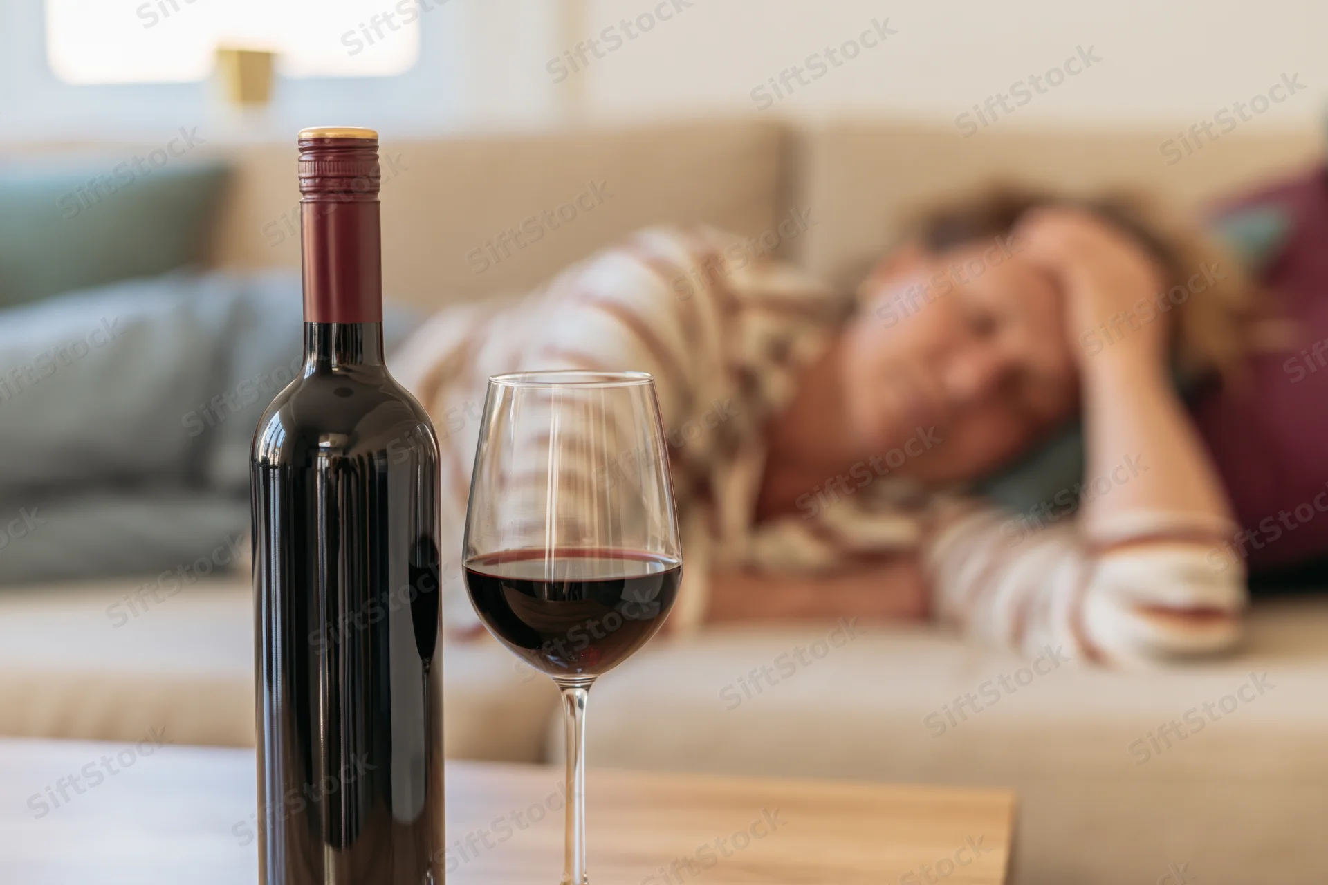 Wine bottle and glass of red wine in focus on table with woman sleeping on couch in background
