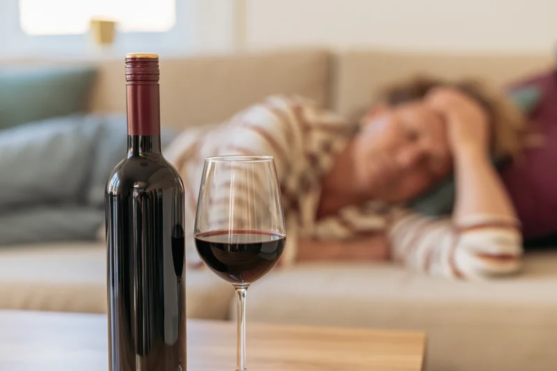 Wine bottle and glass of red wine in focus on table with woman sleeping on couch in background