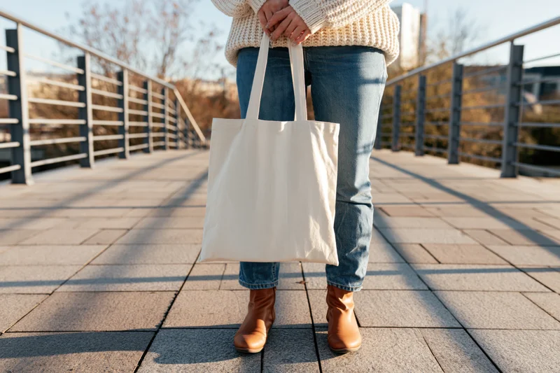 Woman holding blank cream canvas tote bag while walking on a paved bridge outdoors