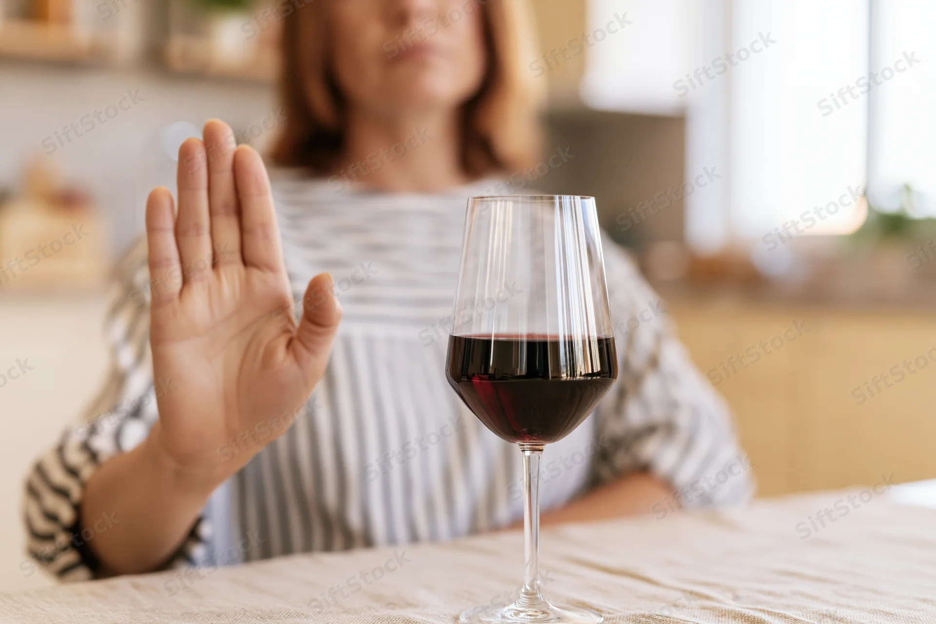 Woman seated at kitchen table raising hand in refusal toward glass of red wine