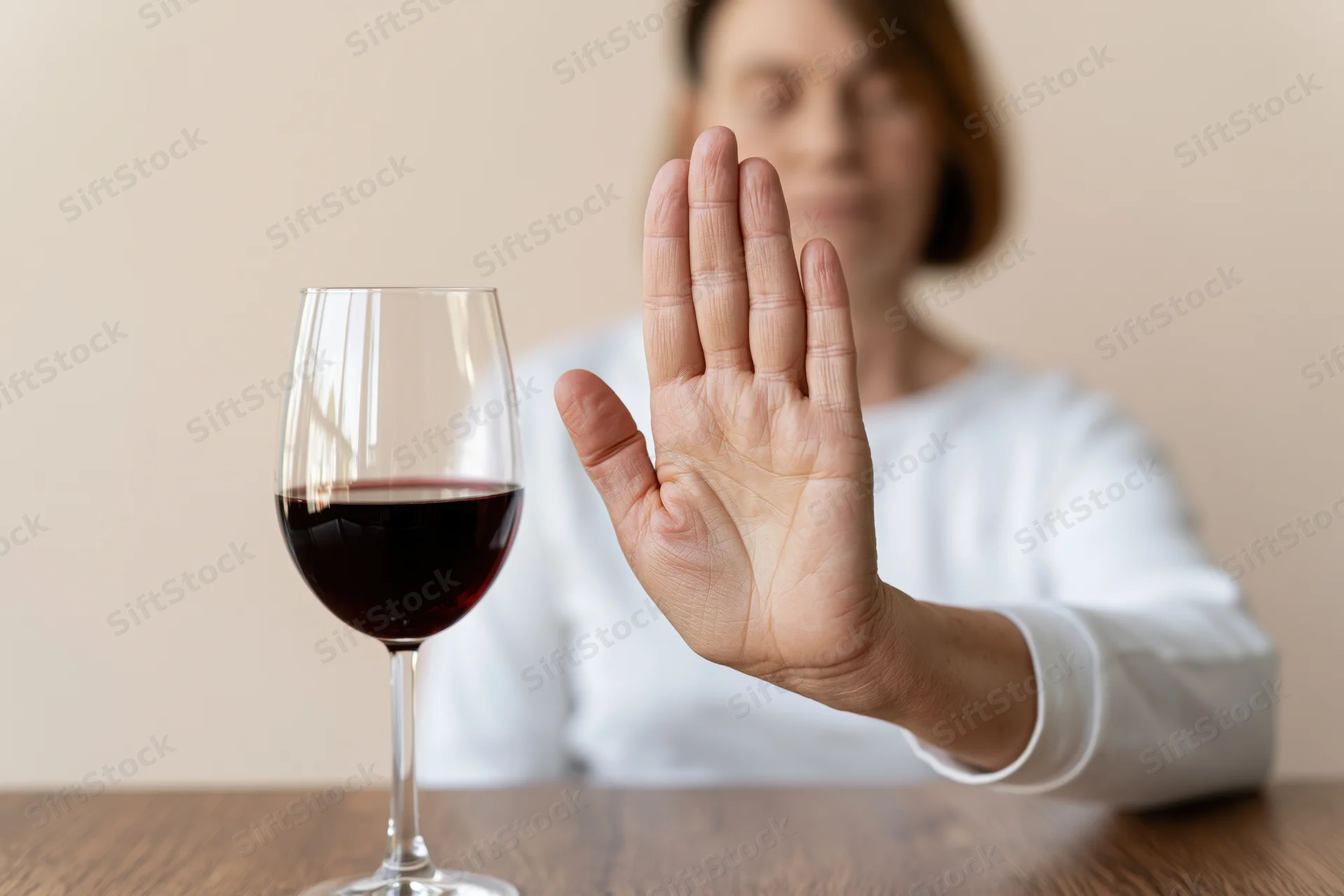 Woman seated at wooden table raising hand to refuse glass of red wine