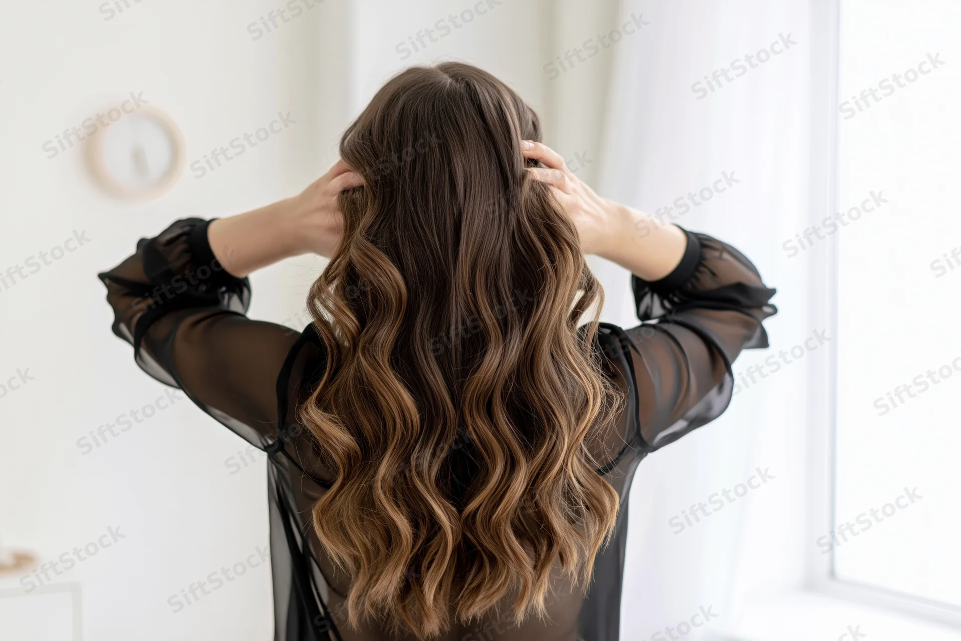 Woman from behind styling her long wavy brown hair with hands in a bright room