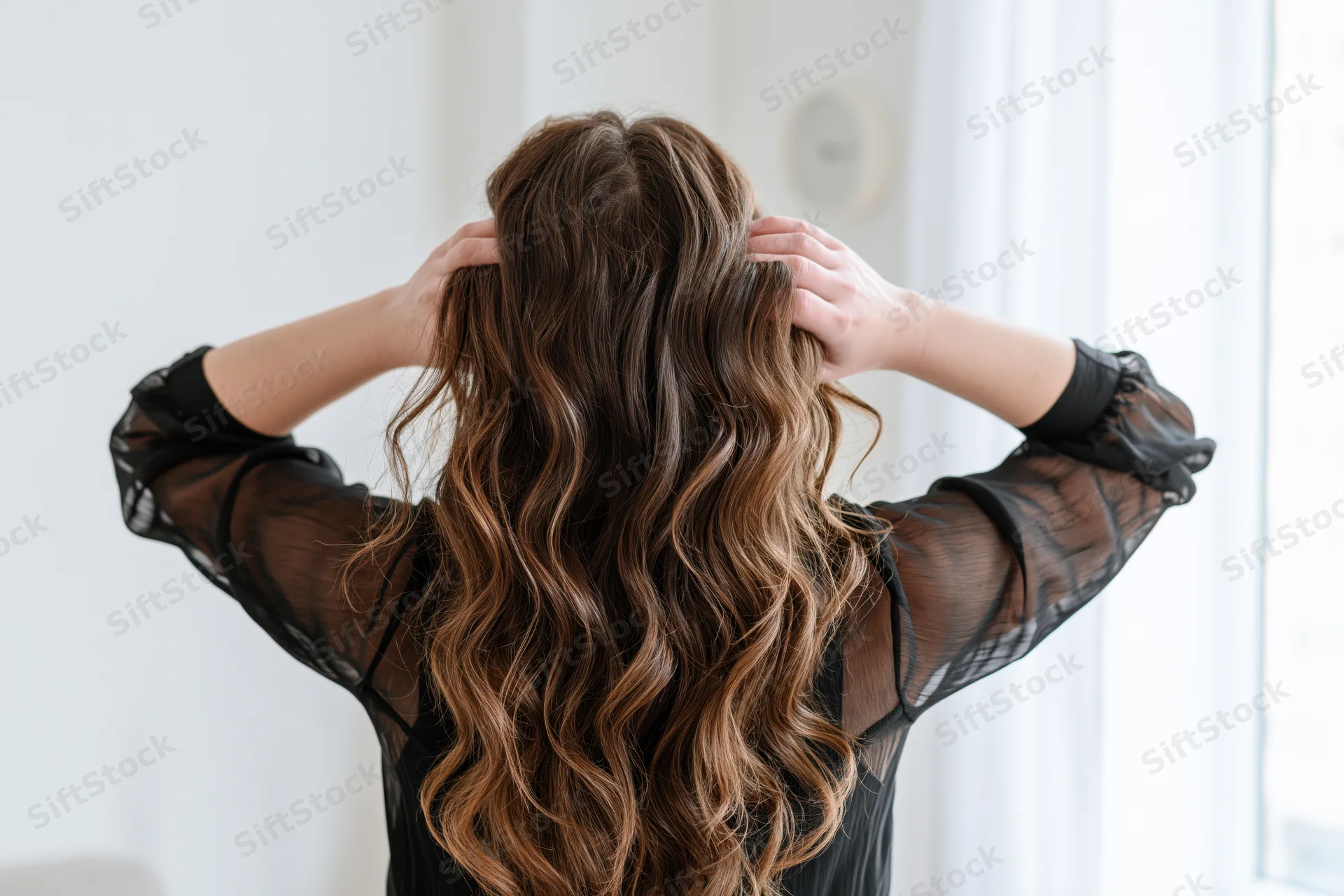 Woman from behind touching her long wavy brown hair with both hands indoors near a window