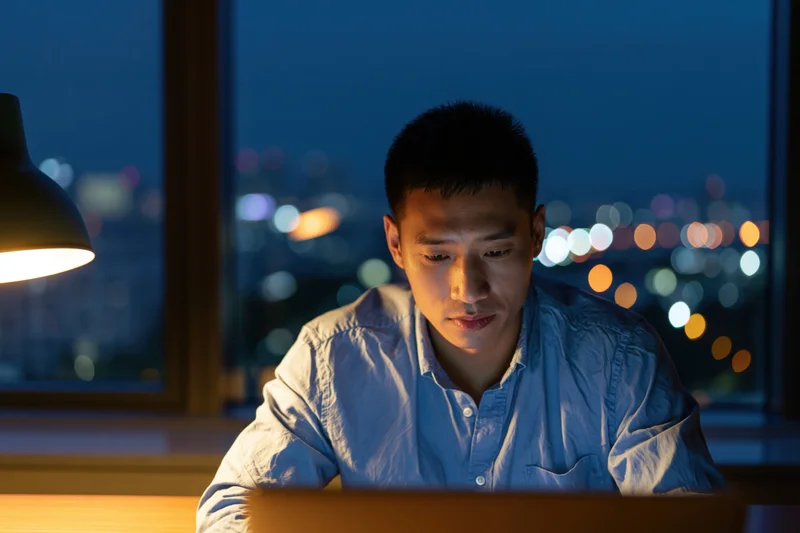 Asian man working on laptop at desk by window at night