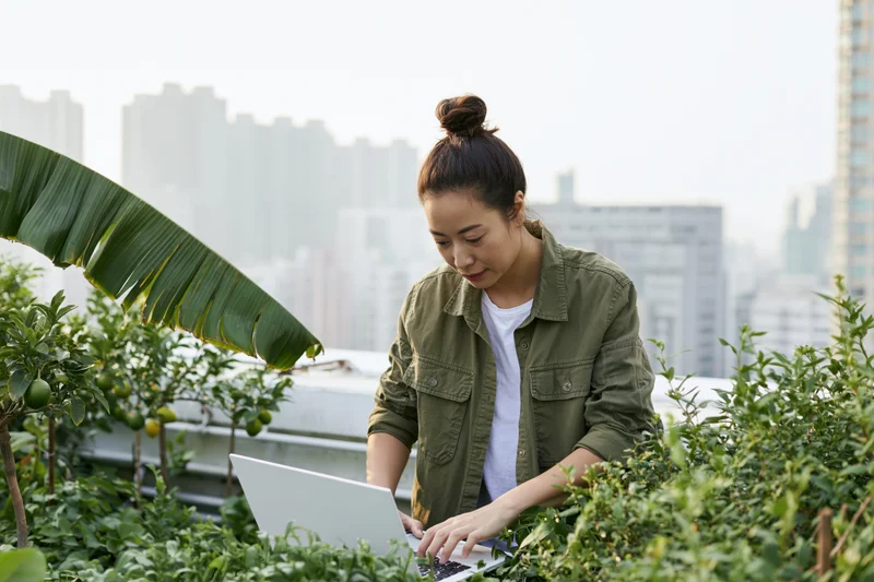 Young asian woman working on laptop among plants on rooftop garden with city skyline in background