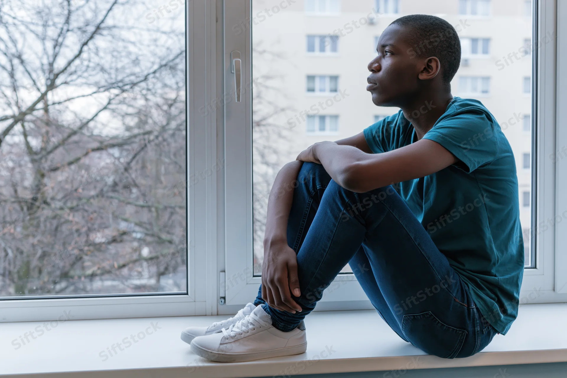 Young black teenage boy sitting on a windowsill looking out at bare trees and apartment buildings