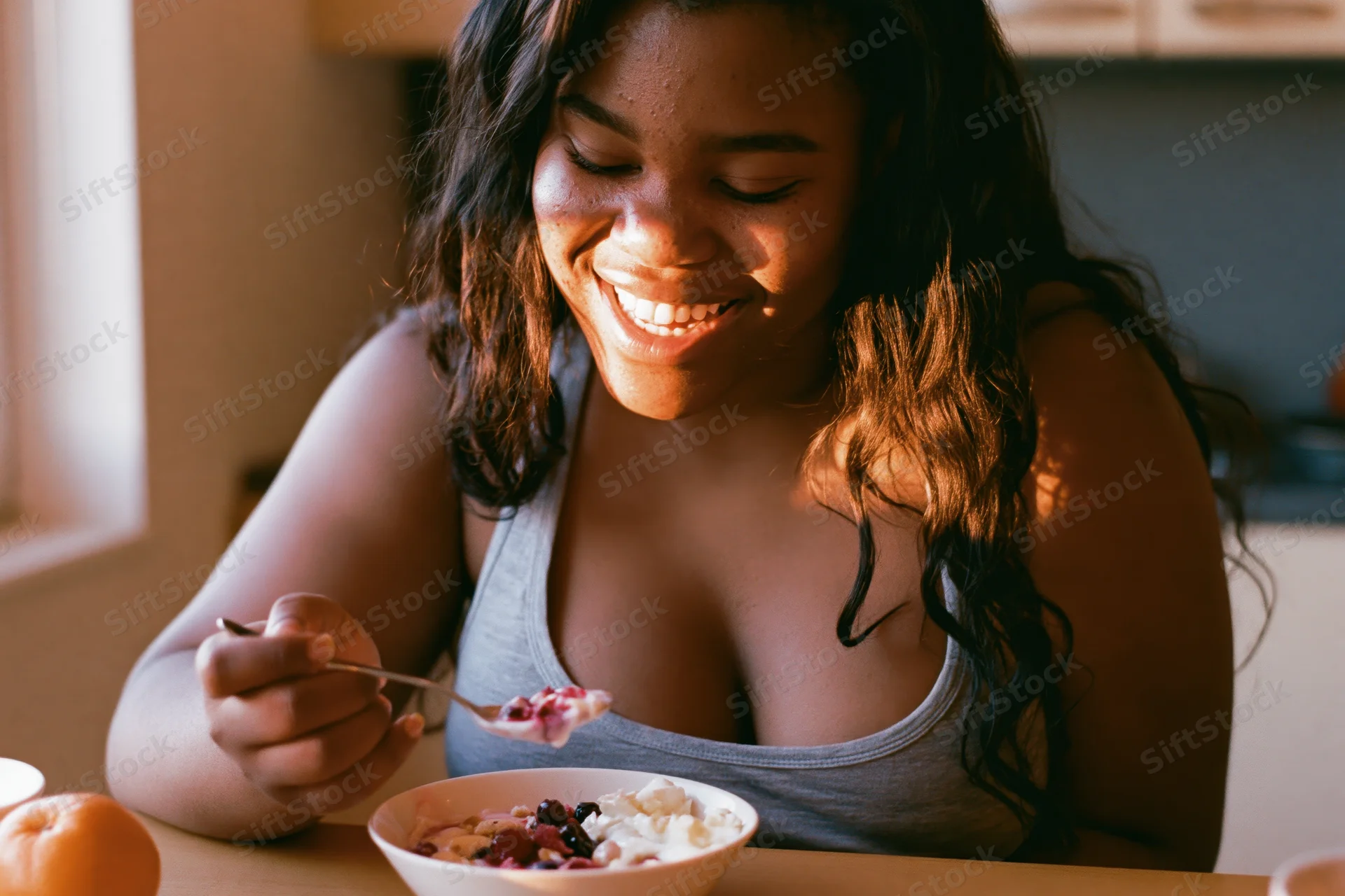 Young black woman smiling while eating yogurt with fruit and granola at a kitchen table in morning sunlight