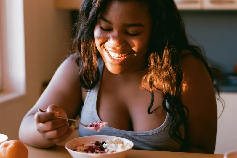 Young black woman smiling while eating yogurt with fruit and granola at a kitchen table in morning sunlight