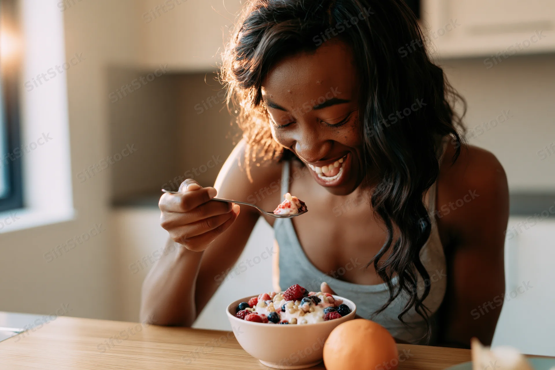 Young black woman smiling while eating yogurt with mixed berries from a bowl at a kitchen table