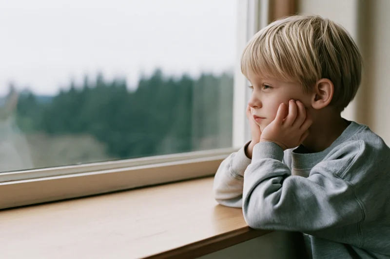 Young blonde boy gazing out window from wooden windowsill indoors
