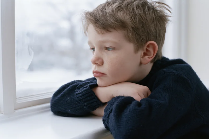 Young boy resting arms on white windowsill gazing out at snowy landscape