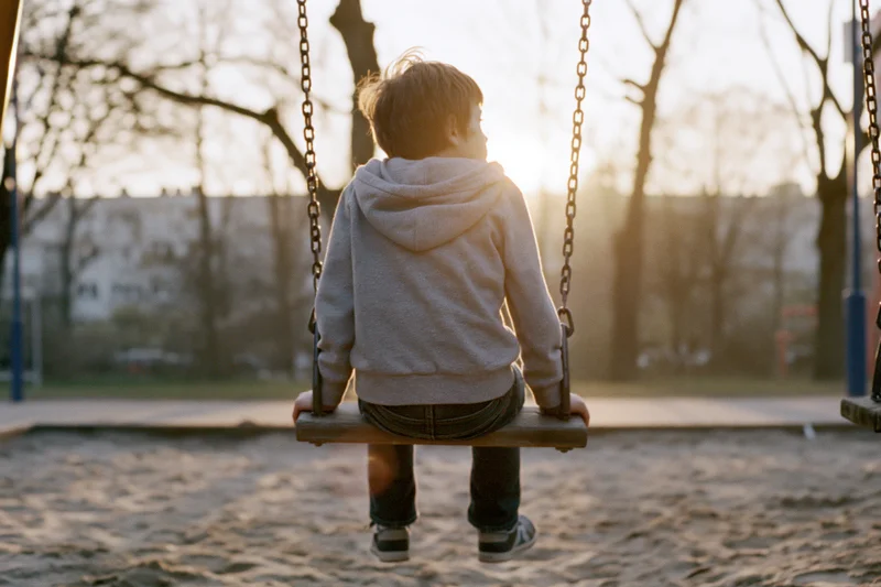 Young boy sitting alone on a wooden swing in a park at sunset