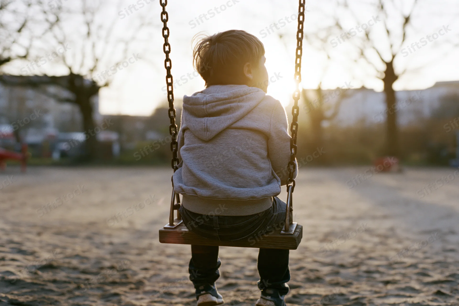 Young boy sitting alone on a wooden swing in a park at sunset