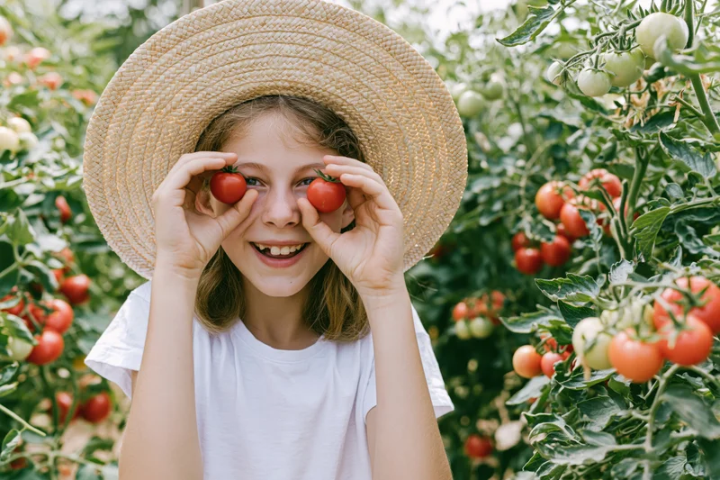 Young girl smiling with tomatoes held over her eyes in a garden