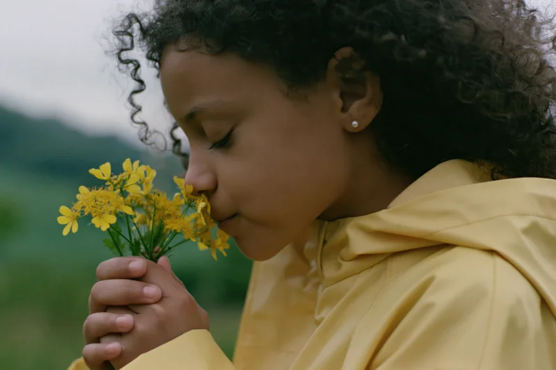 Young girl with curly dark hair smelling a small bunch of yellow wildflowers in a green field