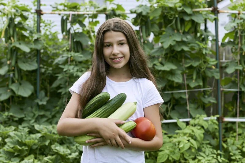 Girl holding zucchini and tomato in a garden with vines and trellis behind her