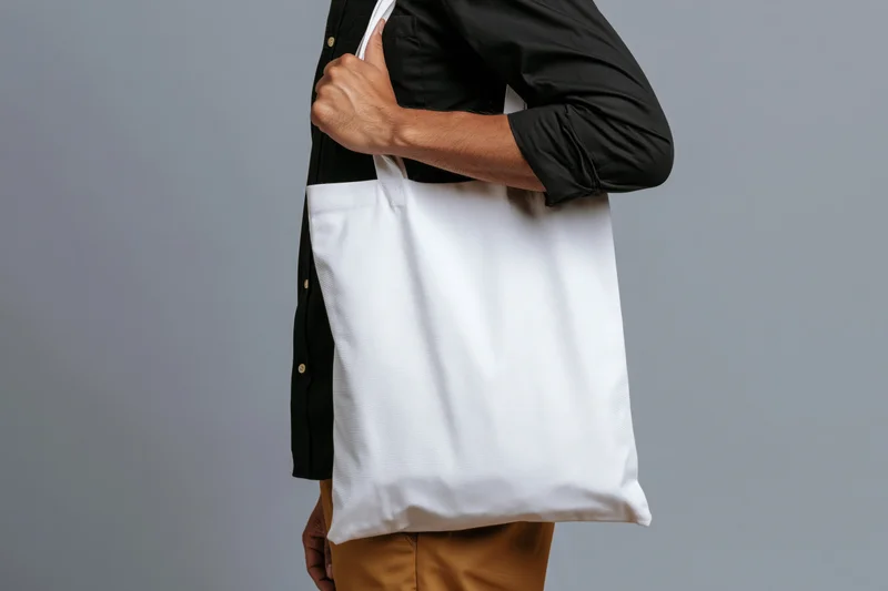 Man holding blank white canvas tote bag over shoulder in studio setting