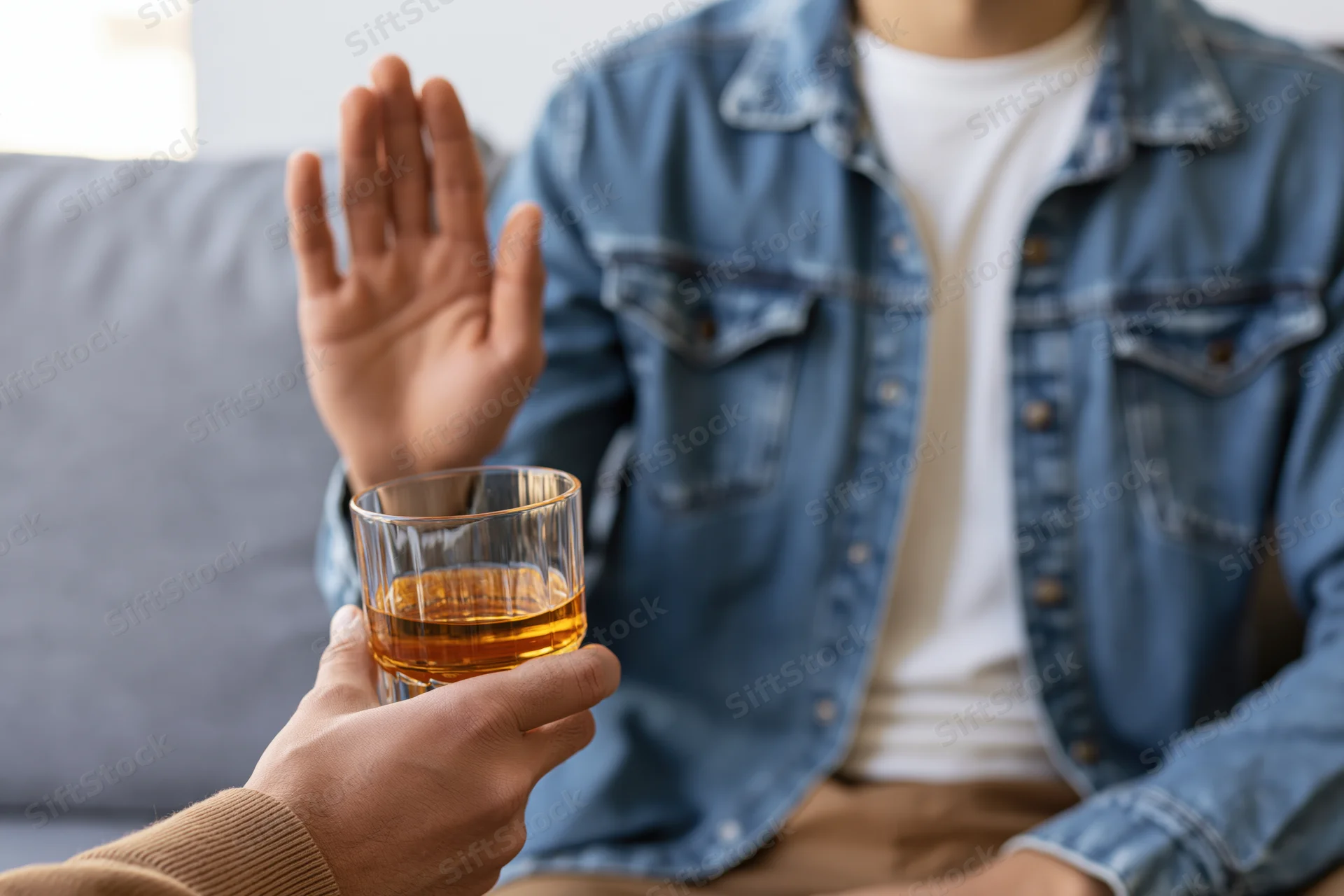 Man in denim jacket seated on couch raising hand to refuse offered glass of whiskey