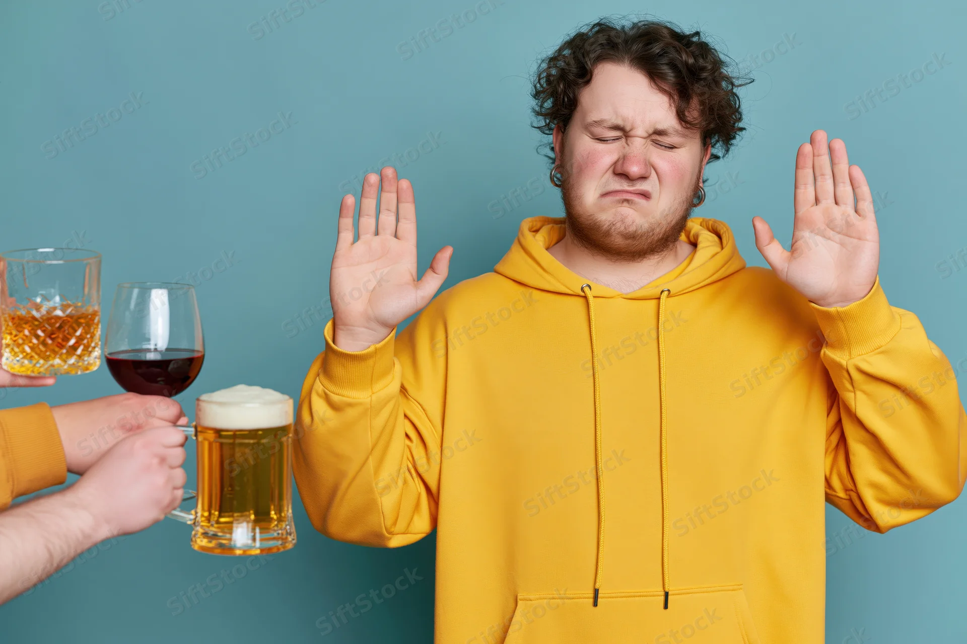 Young man in yellow hoodie raising hands to refuse alcohol offered in glasses