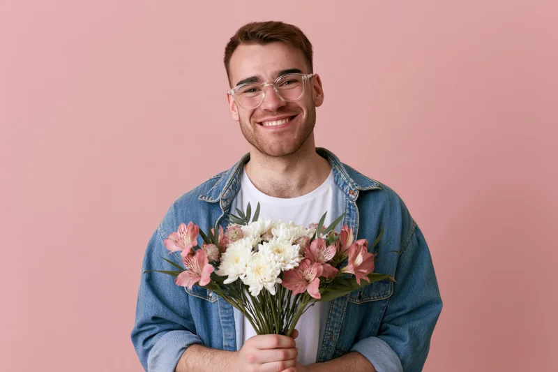 Young man smiling while holding a bouquet of pink and white flowers against a pink background