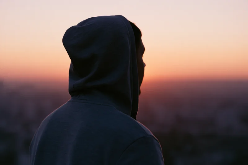 Man in hooded sweatshirt seen from behind overlooking cityscape at sunset