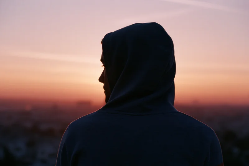 Man in hooded sweatshirt seen from behind silhouetted against sunset cityscape