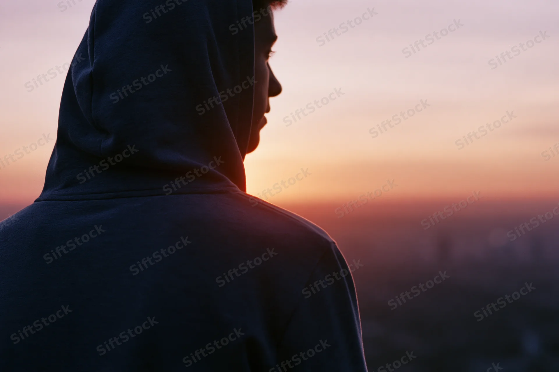 Young man in hoodie seen from behind looking at city skyline at sunset