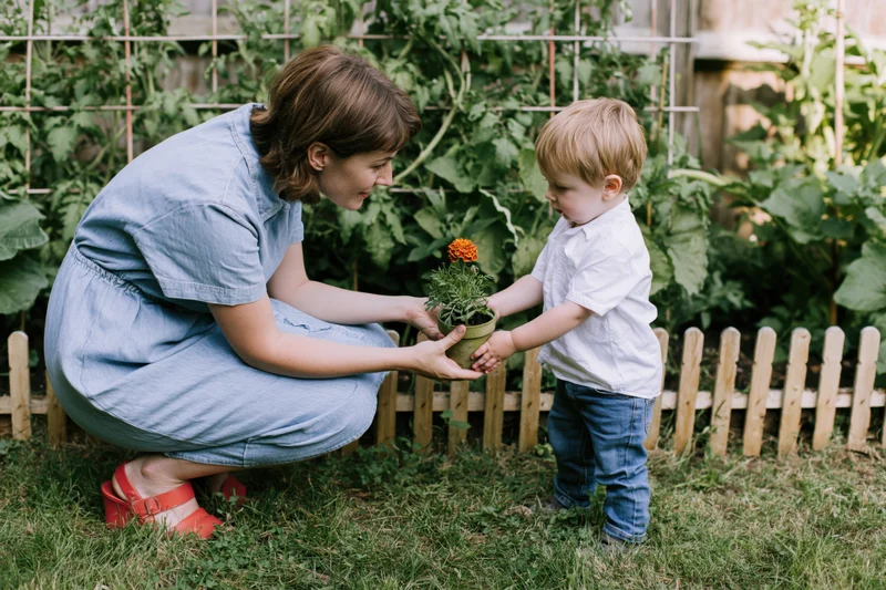 Mother crouching beside toddler son holding potted marigold in backyard garden