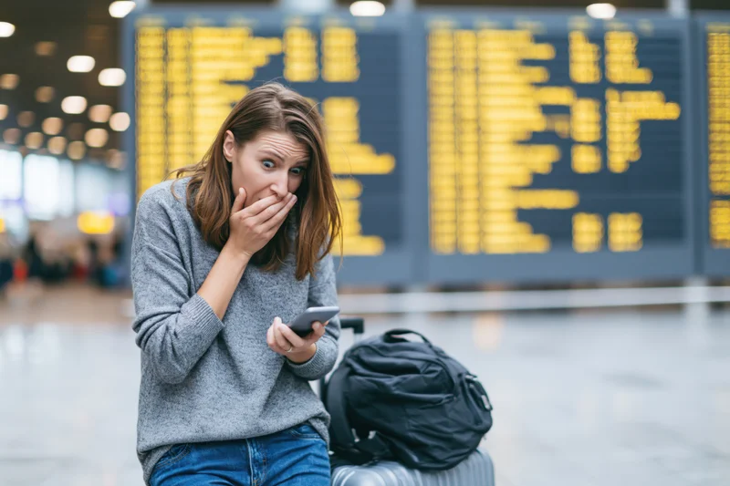 Young woman with shocked expression holding smartphone at airport terminal with luggage and flight display behind her