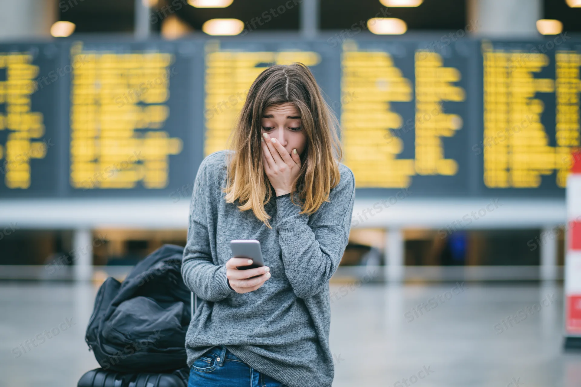 Young woman covering her mouth in shock while looking at smartphone in airport terminal