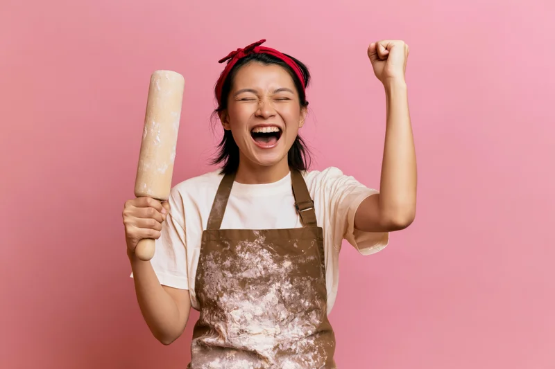 Woman celebrating with clenched fist and rolling pin in apron against pink background