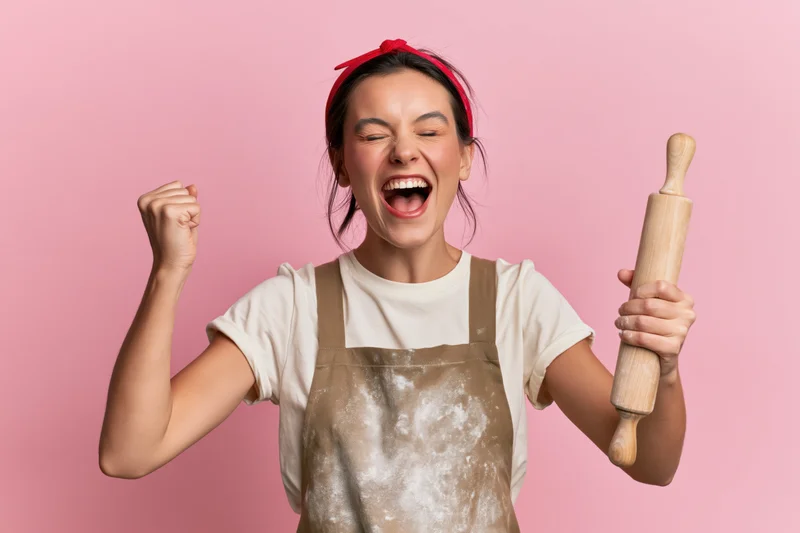 Young woman celebrating with clenched fist while holding wooden rolling pin, wearing flour-dusted apron against pink background